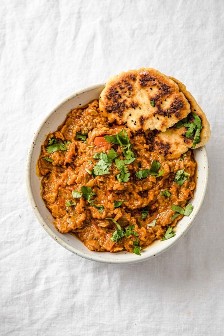 Baingan Bharta (Oven-Roasted Eggplant Curry) in a speckled bowl with naan and garnished cilantro