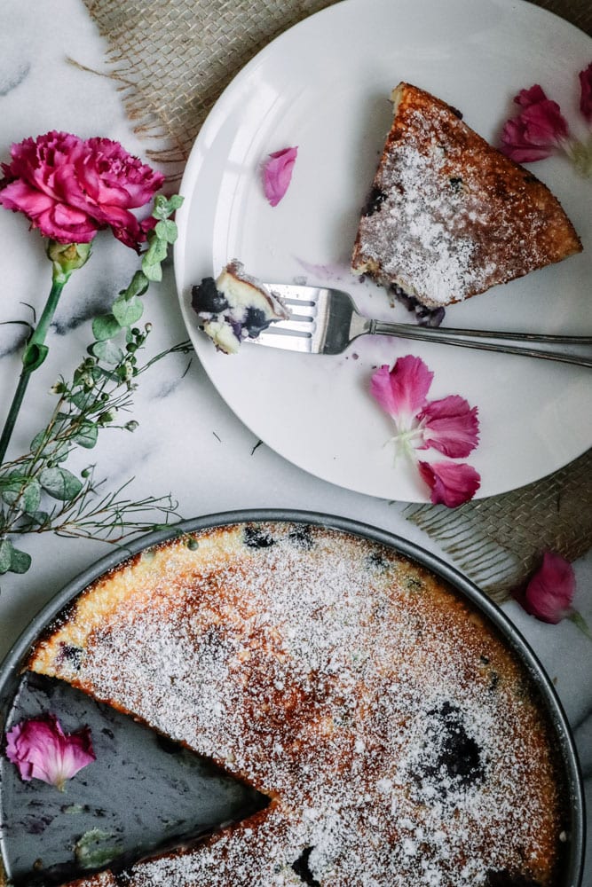 Gluten-Free Blueberry Lemon Clafoutis dusted with powdered confectioners sugar in a baking pan with a sliced removed alongside a partially eaten slice on a plate.