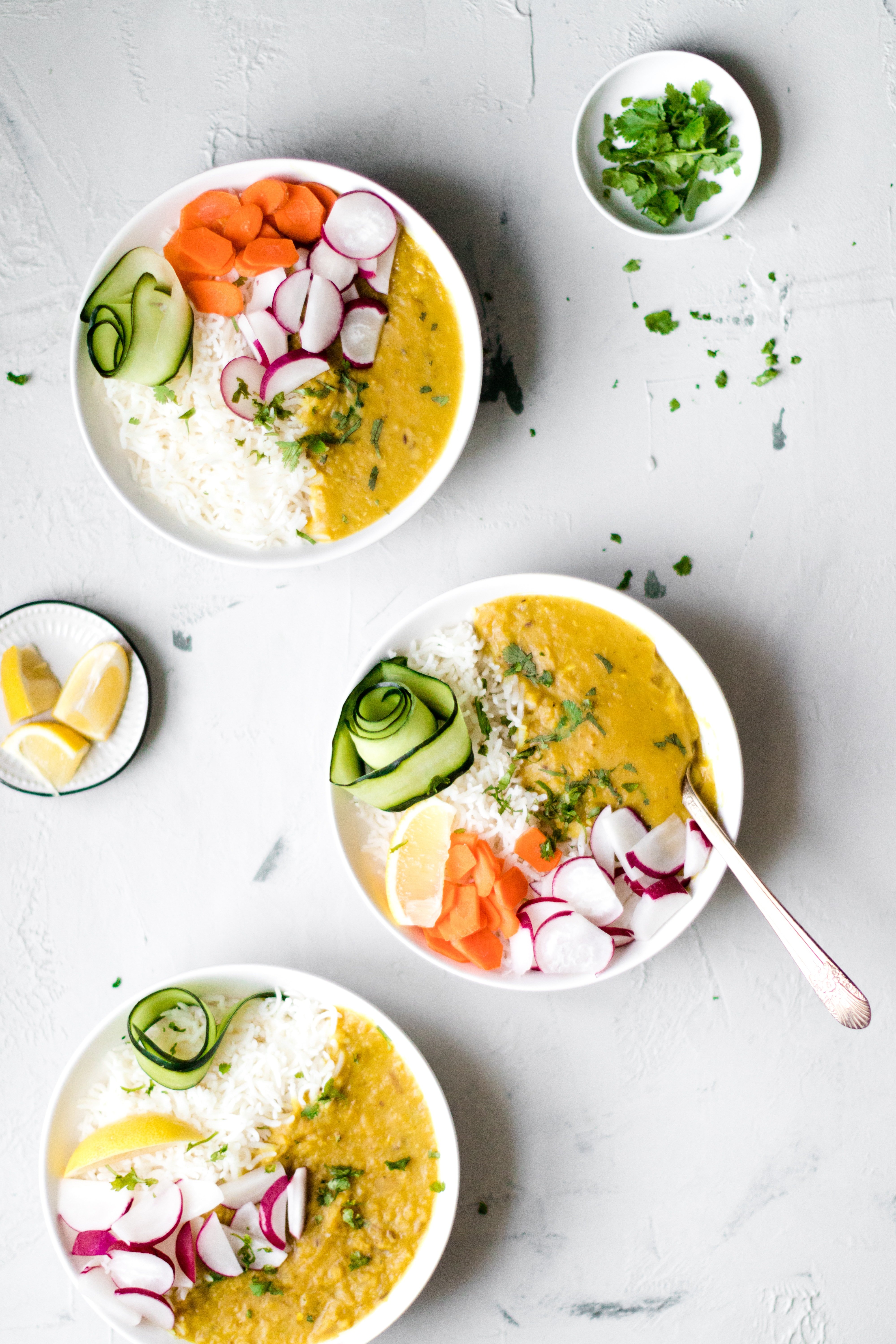 Three white bowls filled with Instant Pot Red Lentil Dal, Basmati rice, ribbons of cucumber, sliced carrots and radishes.