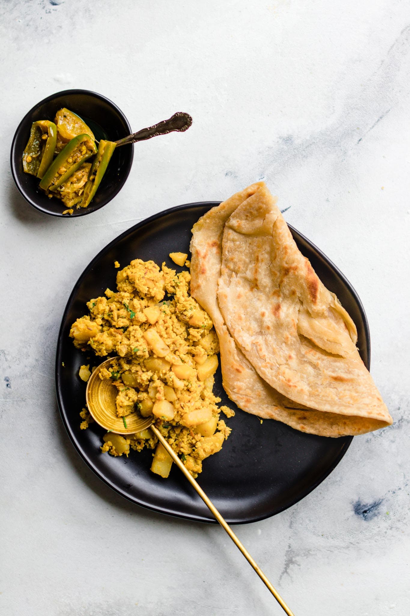 Scrambled Egg and Potato Curry with Paratha on a black plate with a side of achaar (mixed pickle)