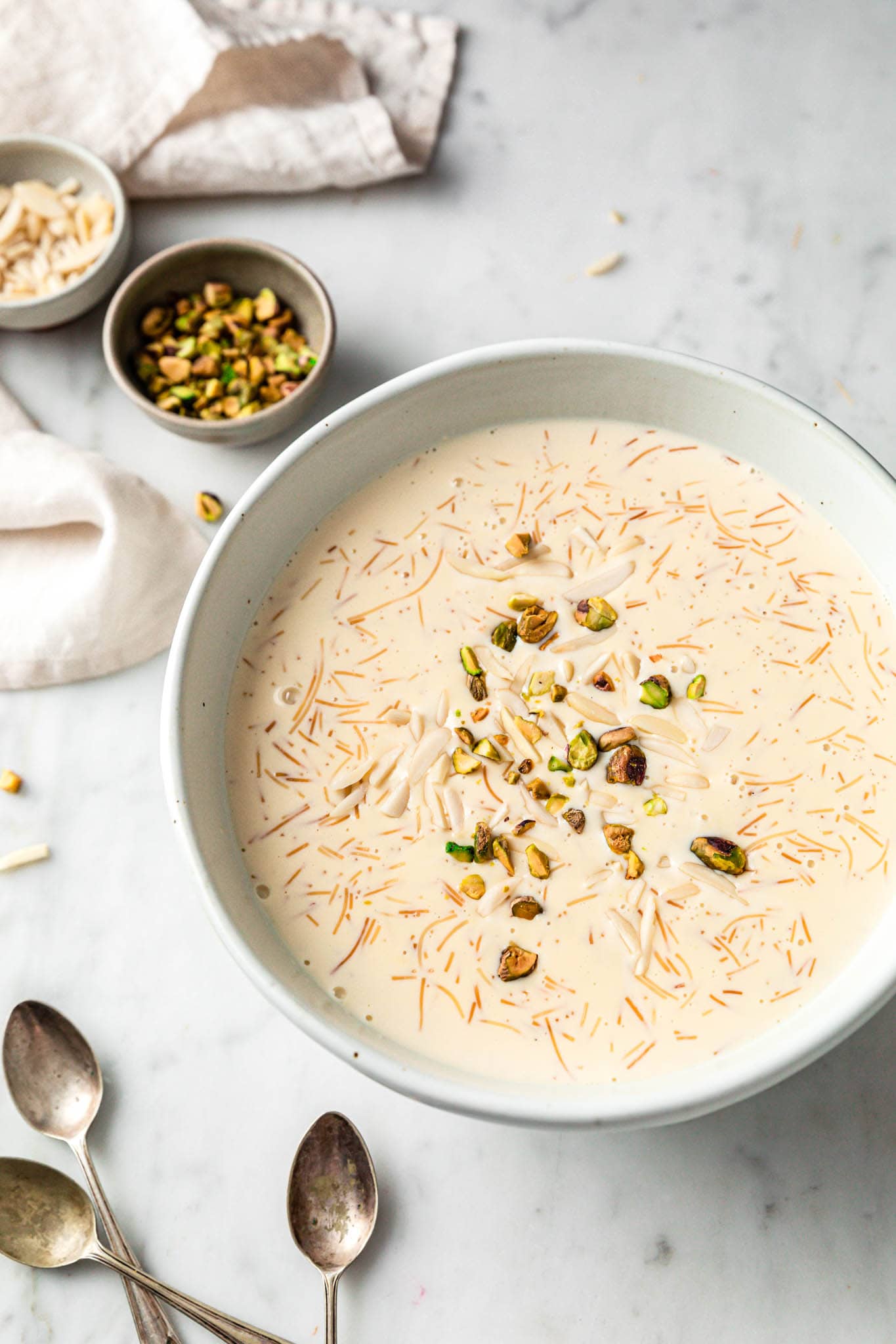 A large bowl with sheer khurma served with spoons and nuts in small bowls