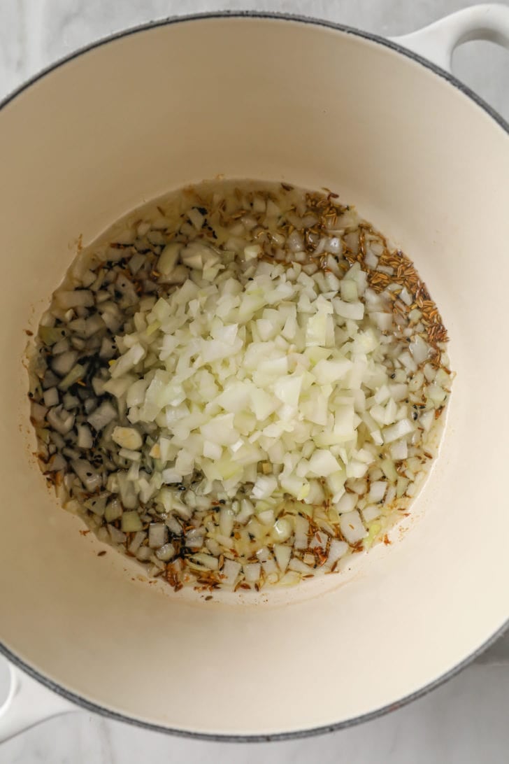 onions added to a hot pan with whole fenugreek seeds, fennel seeds, nigella seeds, and cumin seeds