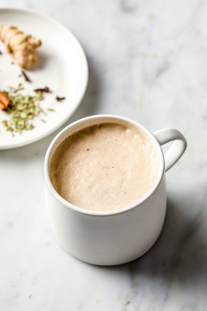 Frothy Masala Chai (Tea) in a white cup with spices in a plate nearby.