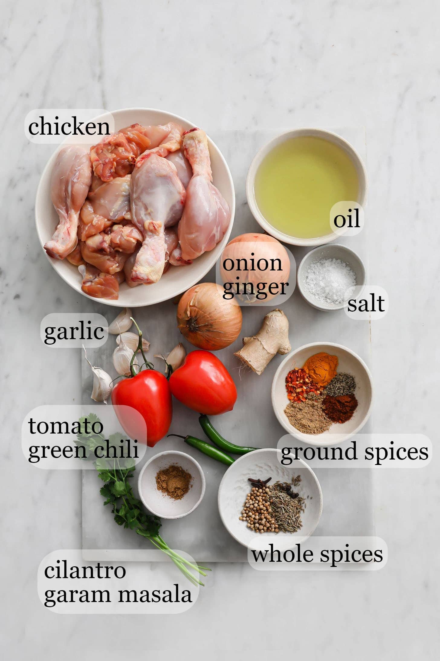 Ingredients for the curry such as chicken, onions, salt, ginger, garlic, tomatoes, green chili peppers, and spices on a marbled surface