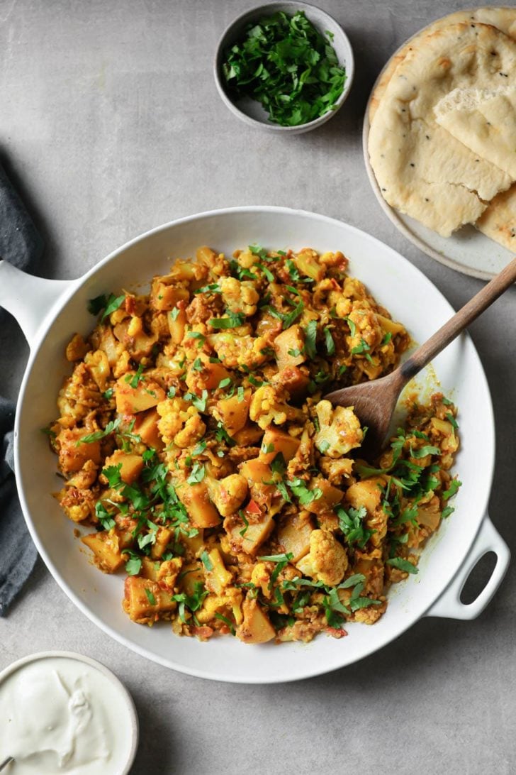 Cauliflower and potato curry garnished with cilantro in a white skillet with a wooden spoon