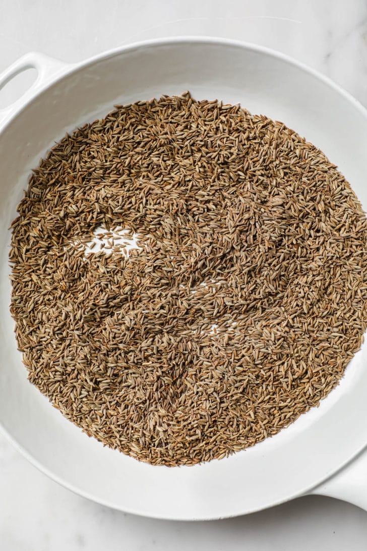 Toasting cumin seeds in a white skillet.