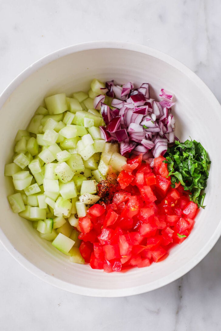 Diced cucumber, tomato, red onion, cilantro with some spice in a white bowl.