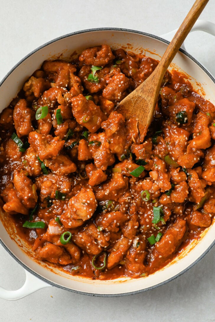Manchurian chicken in a pot with a wooden spoon and garnished with sesame seeds and green onion