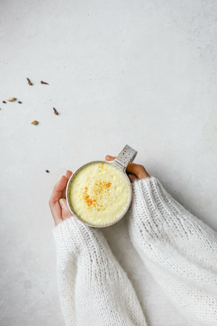 Holding a speckled mug filled with Turmeric Milk with some turmeric powder dusted on top.