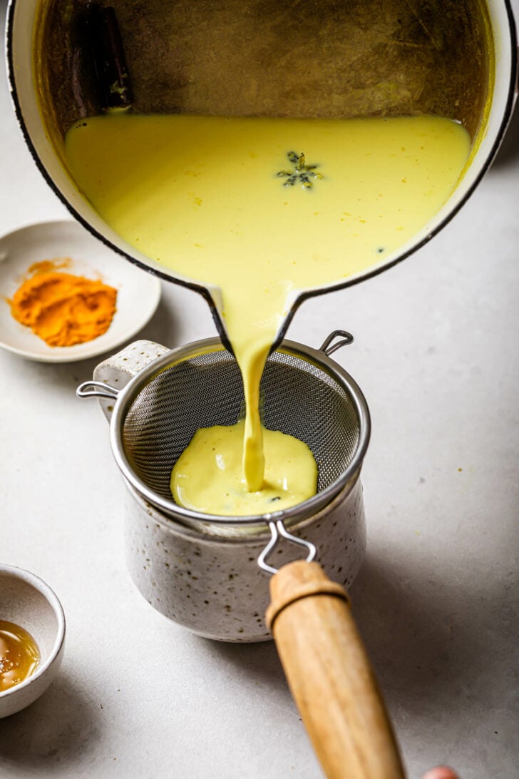Pouring Golden Milk into a speckled mug through a strainer.
