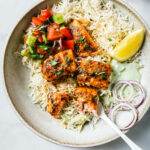 Top view of a partially eaten plate of Salmon Tikka with rice, vegetables, onion and lemon.