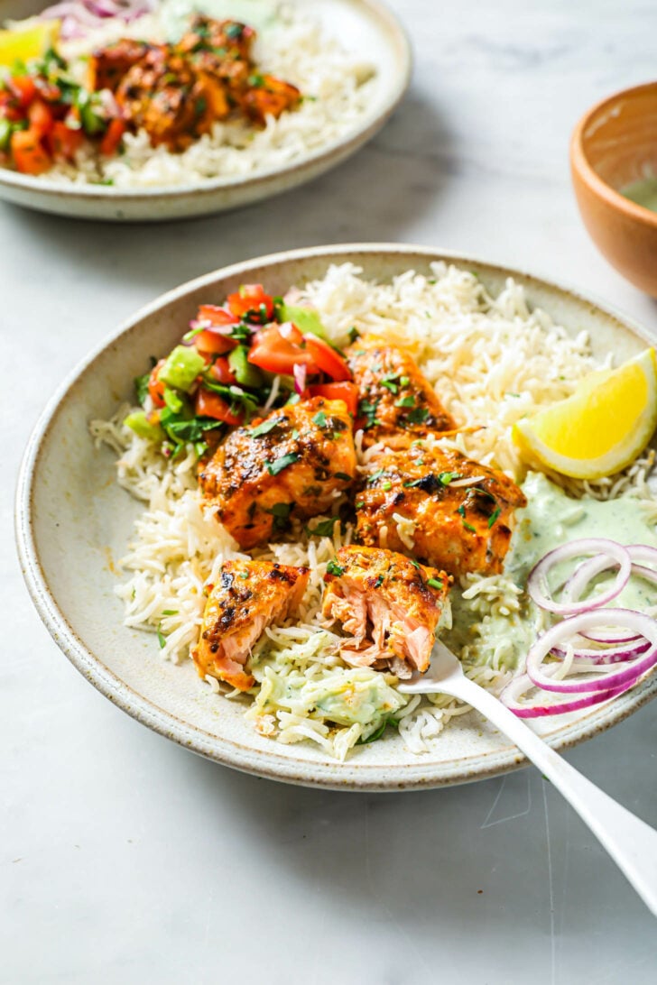 Side view of partially eaten plate of Salmon Tikka with rice, vegetables, onion and lemon.