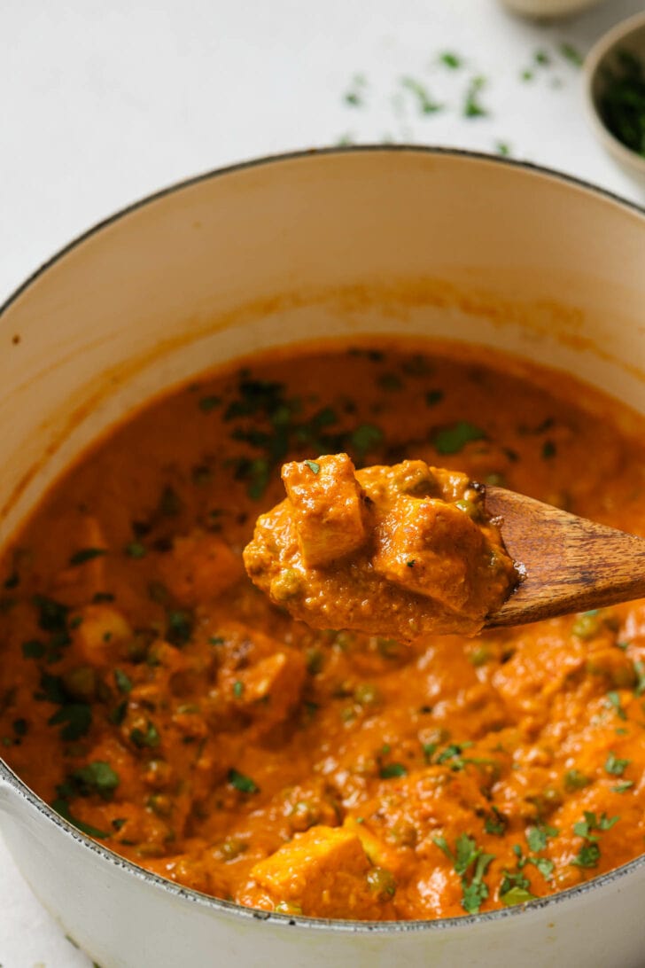 Holding a spoonful of saucy Paneer cubes over a dutch oven.
