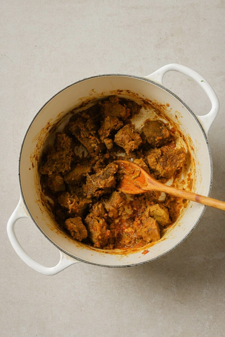 Cooked goat/lamb in a dutch oven with a wooden spoon.