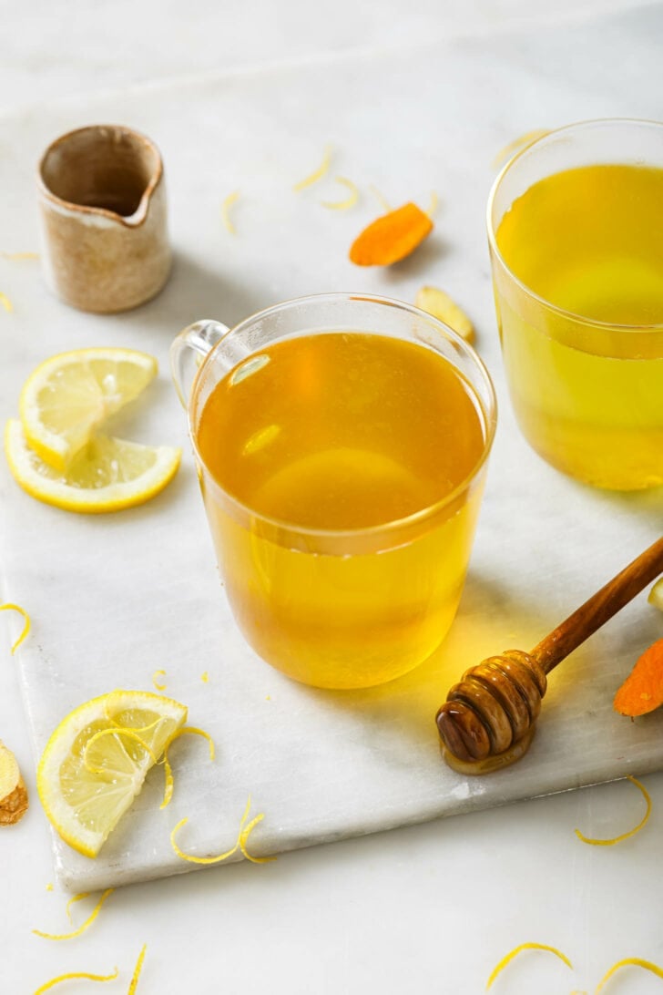 Two mugs of Turmeric Tea on a wooden surface with slices of lemon, turmeric and a honey dipper.