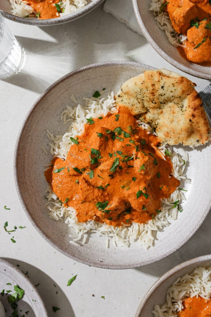 A bowl of rice topped with Butter Chicken with some naan on the side.