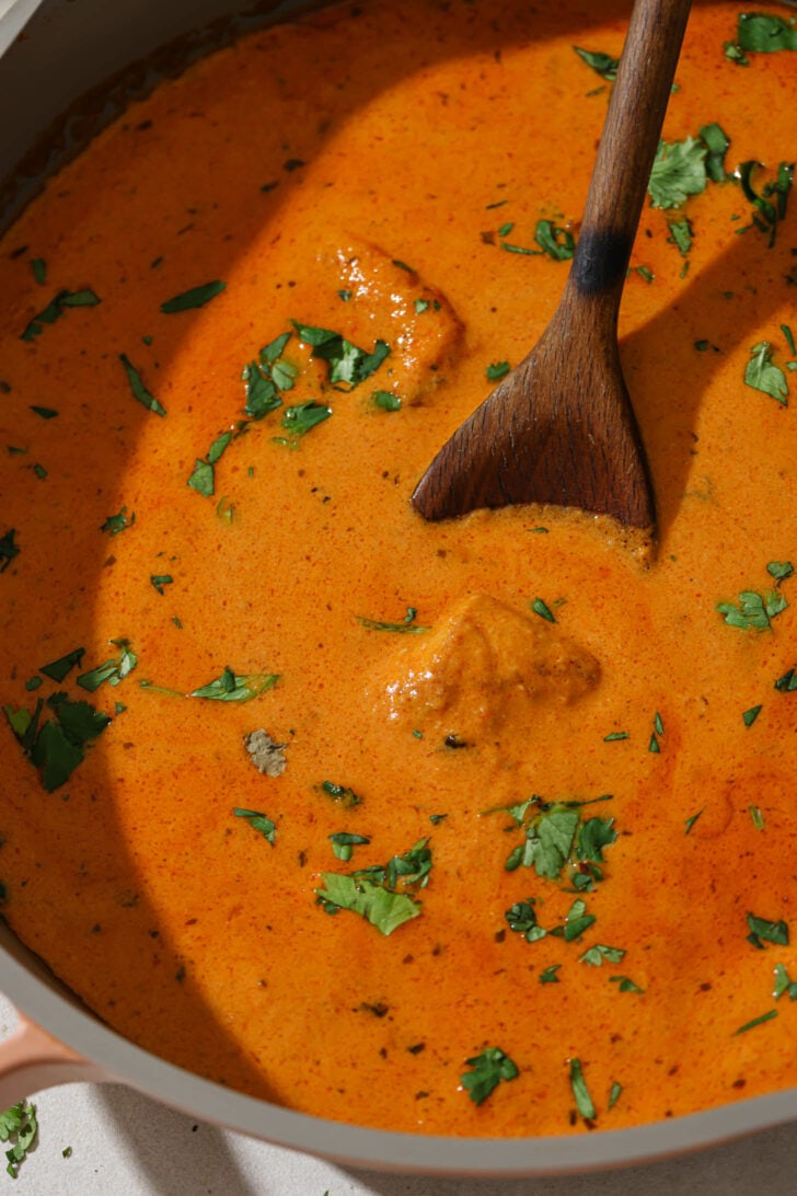 Close up of Butter Chicken in a skillet garnished with cilantro. with a wooden spoon.