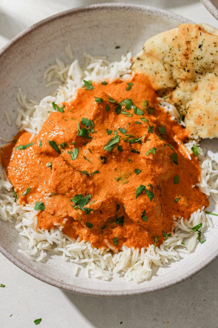 A bowl of rice topped with Butter Chicken with some naan on the side.