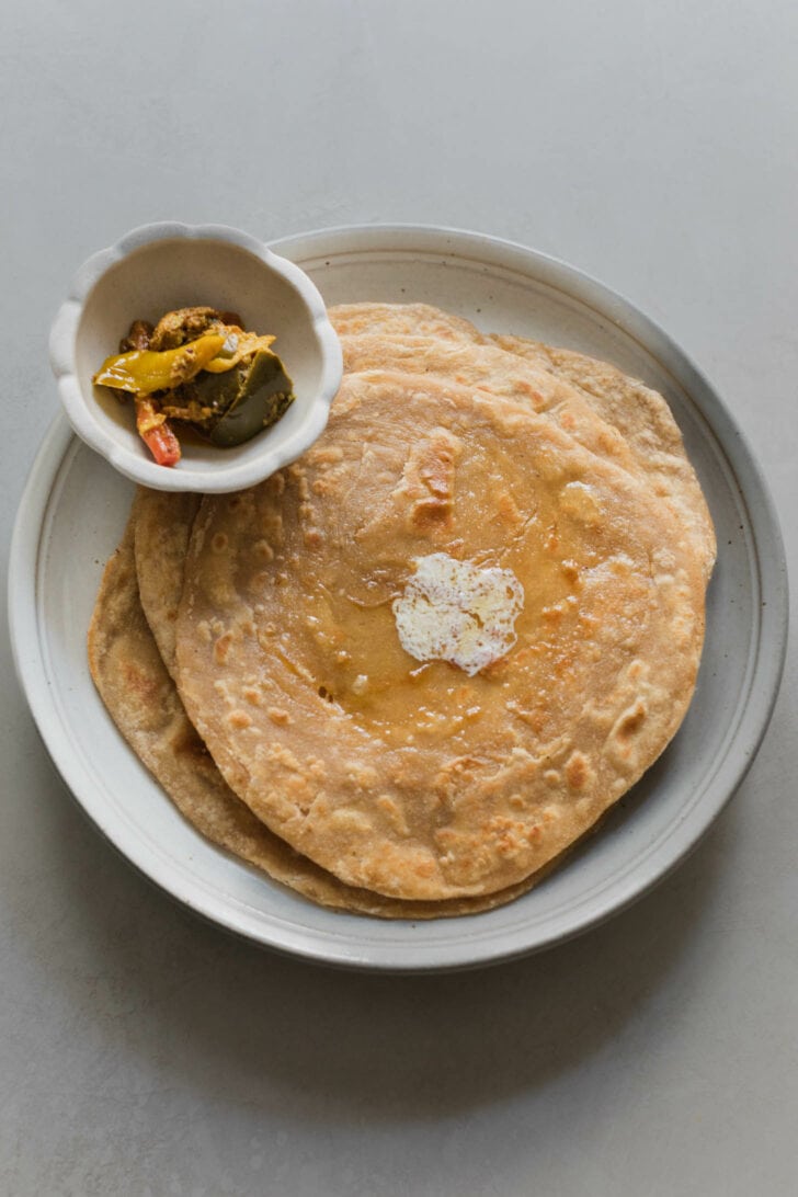 A stack of Laccha parathas with a dollop of butter that has melted on top alongside a bowl of achaar.