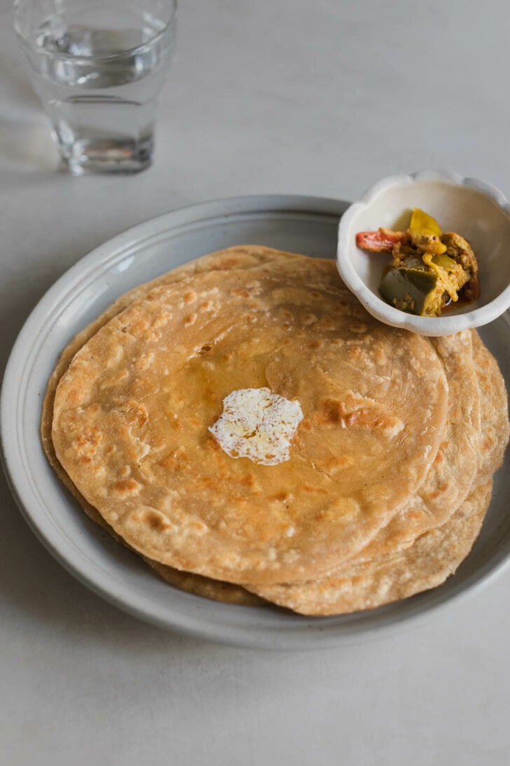 A side view of stack of laccha paratha with a dollop of butter that has melted on top alongside a bowl of achaar.