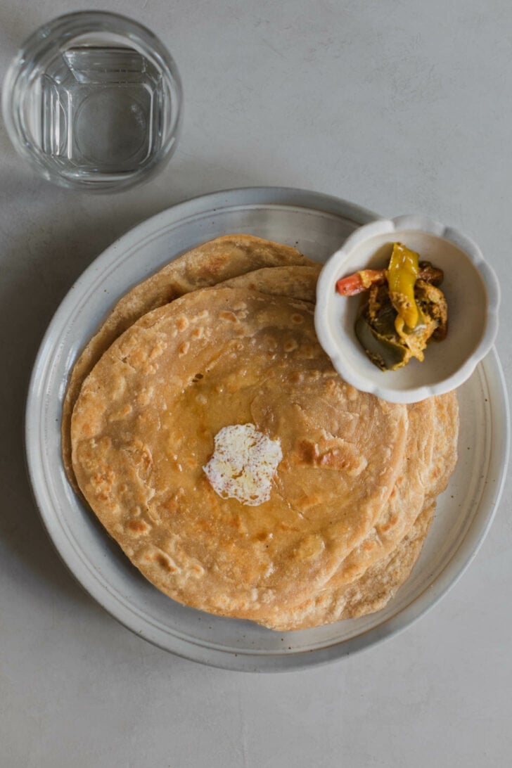 A stack of paratha with a dollop of butter that has melted on top alongside a bowl of achaar.