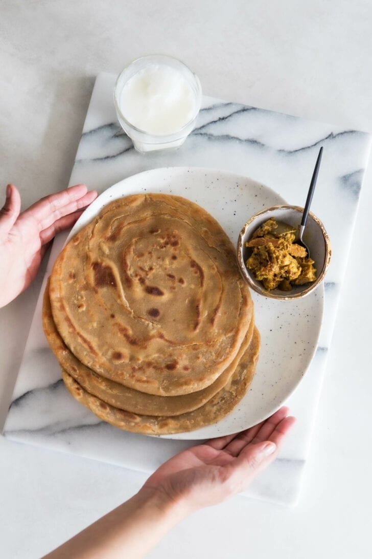 Holding a plate of three lachha parathas with a bowl of achaar on the side.